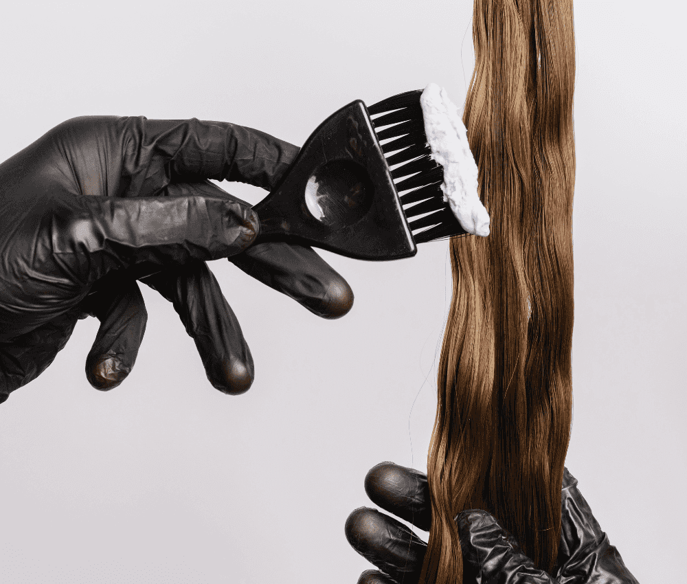 Gloved hand applying hair dye to long brown hair with a brush.
